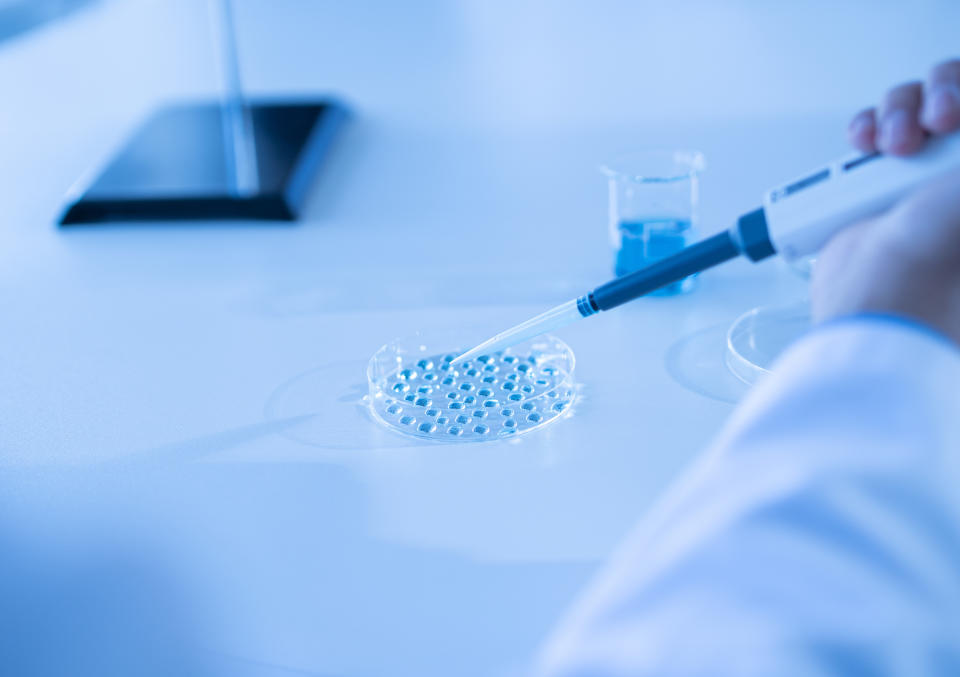 Researcher using a pipette to drop liquid into a multiwell plate, with a tablet in background, in a lab setting