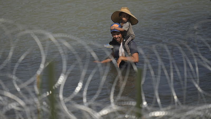 Migrants, one a child, stand in the Rio Grande behind concertina wire as they try to enter the U.S. from Mexico near the site where workers are assembling large buoys to be used as a border barrier in Eagle Pass, Texas, on July 11, 2023. The floating barrier is being deployed in an effort to block migrants from entering Texas from Mexico.