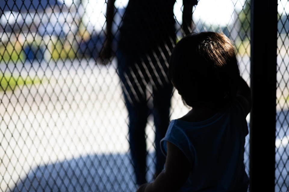 Child standing near a chain-link fence, reaching towards an adult figure in the background. Faces are not visible