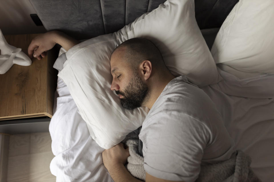 A bearded man with a shaved head sleeps peacefully on a white pillow in a cozy bed. He wears a white t-shirt and has a calm expression