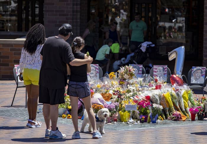 One person puts their arm around another's shoulders as they stand near a collection of flowers and portraits