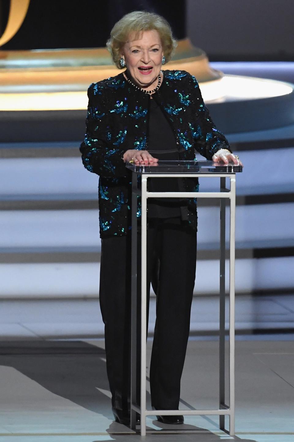 Betty White at an awards event, wearing a sparkling outfit and standing at a podium with 