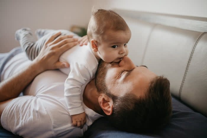 A parent lying on the couch and kissing their young baby lying on top of them.