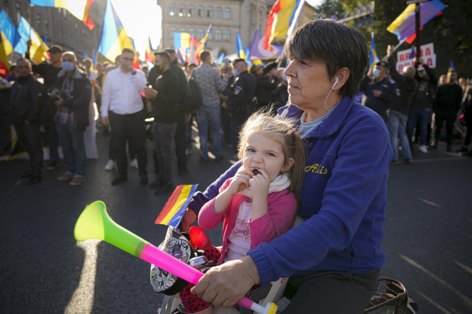 A girl sitting on a bicycle rides by protesters waving flags during an anti-government protest organised by the far-right Alliance for the Unity of Romanians or AUR, in Bucharest, Romania, Saturday, Oct. 2, 2021. Thousands took to the streets calling for the government's resignation, as Romania reported 12,590 new COVID-19 infections in the past 24 hour interval, the highest ever daily number since the start of the pandemic. (AP Photo/Vadim Ghirda)