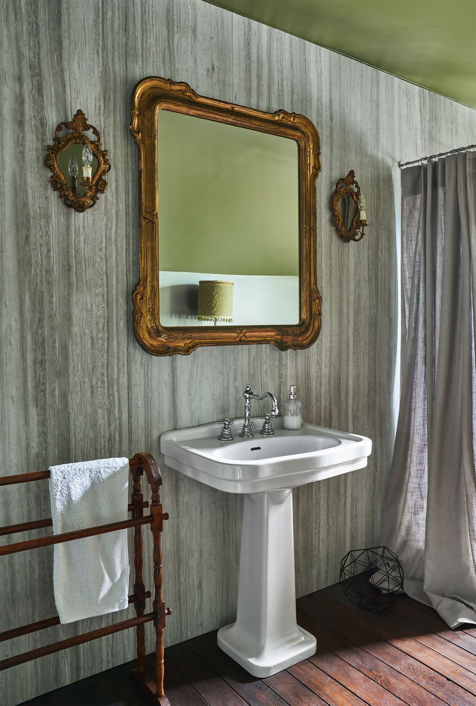 in a bathroom the walls are travertine covered, wooden drying rack with a towel, pedestal sink with silver fittings, antique mirror and two sconces above sink, linen shower curtain, slat wood floor, green ceiling