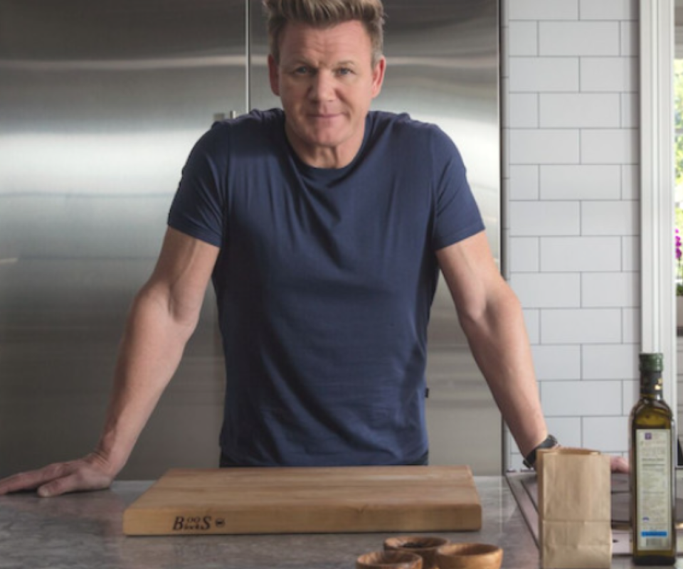 Gordon Ramsay stands in a kitchen with his hands on a grey surface with a wooden chopping block in front of him.