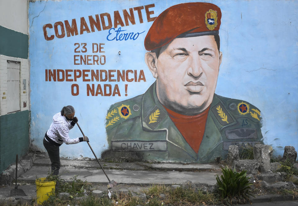 Una mujer porta una mascarilla mientras limpia una acera frente a un mural del fallecido presidente venezolano Hugo Chávez durante una jornada de limpieza y desinfección organizada por el consejo comunal y organización colectiva del vecindario 23 de Enero de Caracas, Venezuela, el miércoles 29 de abril de 2020. (AP Foto/Matias Delacroix)