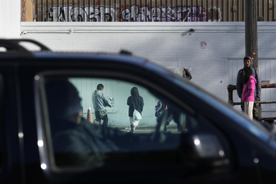 A motorist drives past immigrants standing outside and returning to a shelter in the Pilsen neighborhood of Chicago, Tuesday, Dec. 19, 2023. The death of a 5-year-old migrant boy and reported illnesses in other children living at the shelter has raised concerns about the living conditions and medical care provided for asylum-seekers arriving in Chicago. Four more people living in the same shelter — mostly children — were hospitalized with fevers this week. (AP Photo/Charles Rex Arbogast)