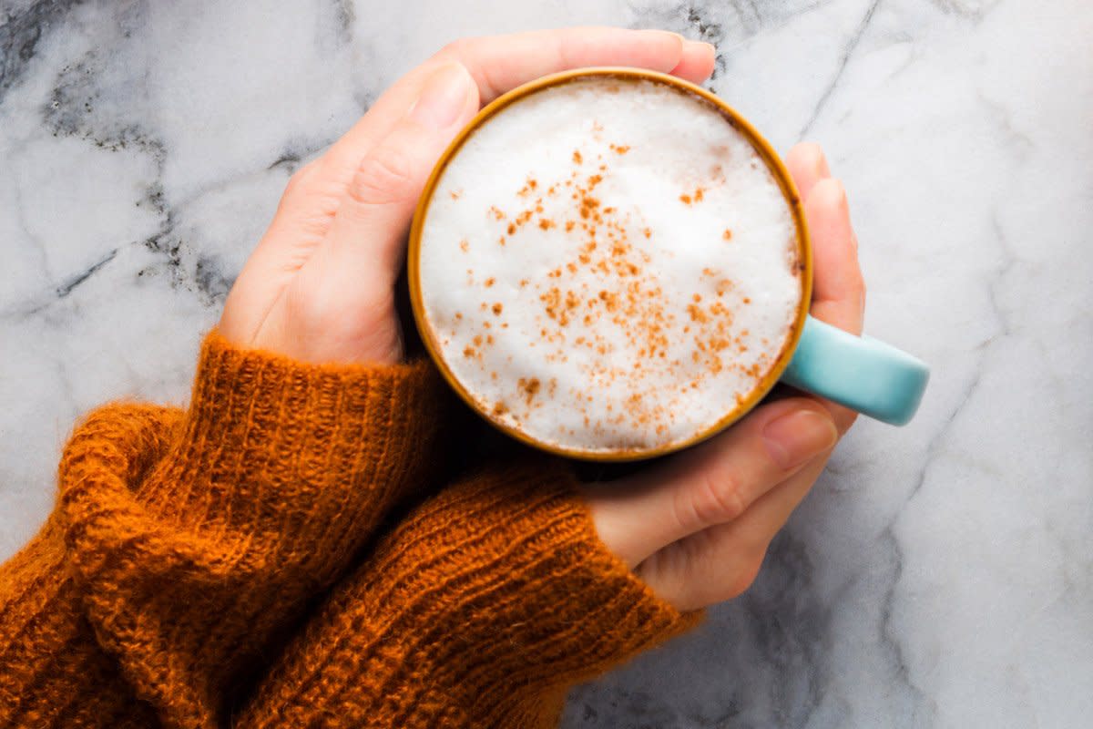 A woman wearing an orange sweater has her hands around a coffee mug with a pumpkin spice latte inside of it. 