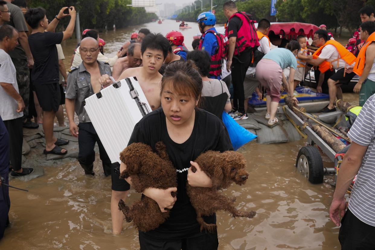A woman carries two dogs in her arms while standing in ankle-deep water among dozens of other people and rescue workers..