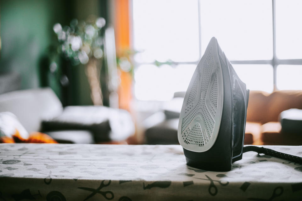 An iron placed upright on an ironing board with a blurred home background