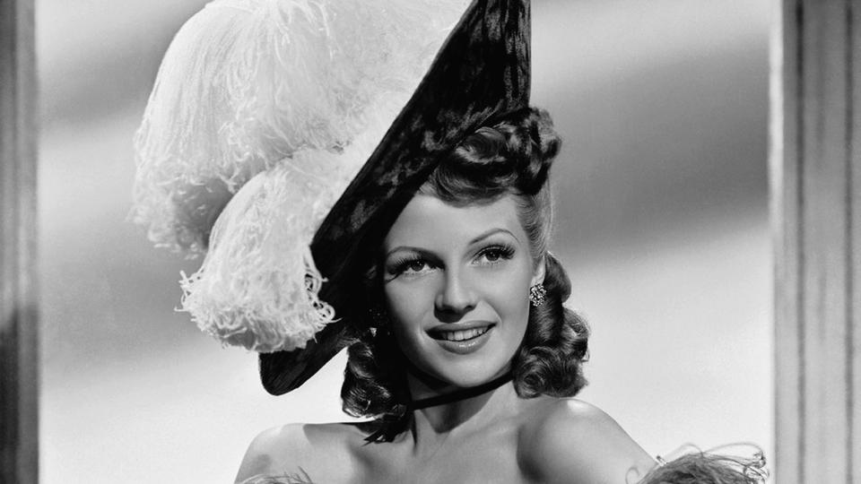 rita hayworth poses with one hand on her hip, she wears a sequin gown with a large feather trim, a matching hat and white gloves