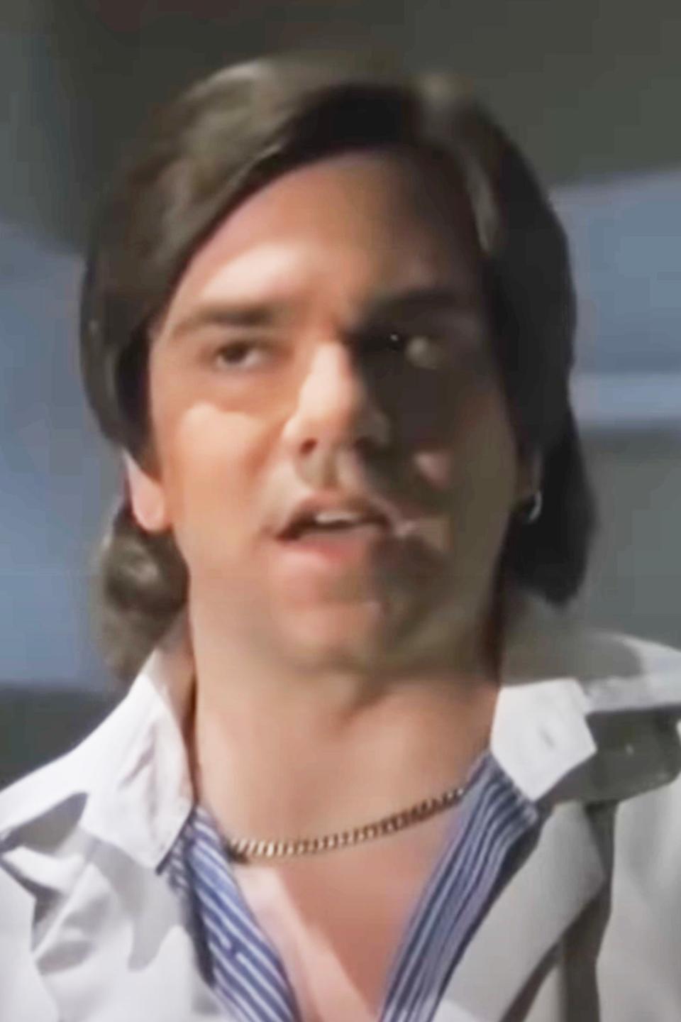 A young Matt Berry with medium-length dark hair is wearing a white shirt over a striped top and a chain necklace