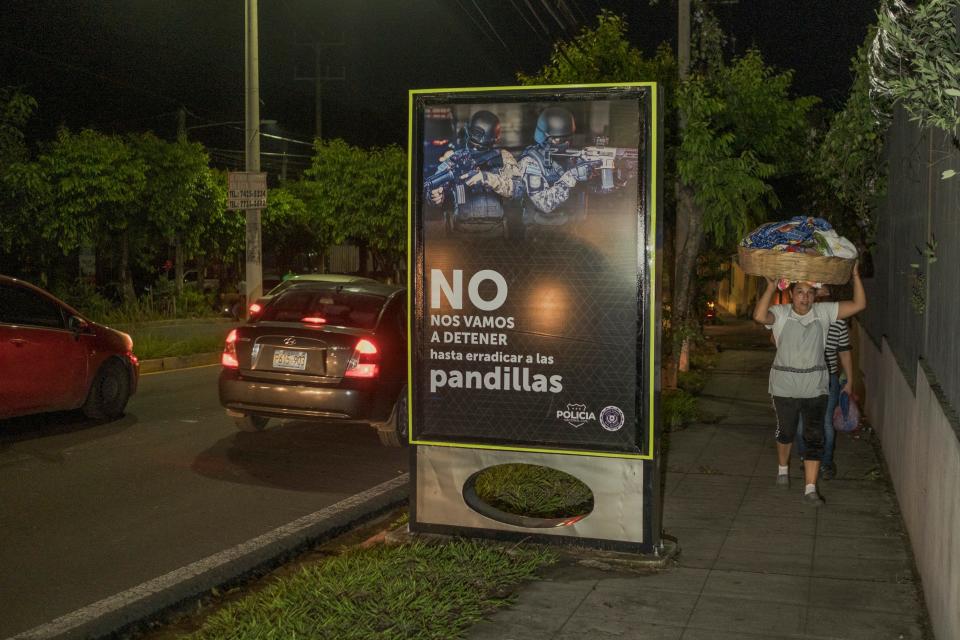 Women are seen carrying things, at night, past a sign with images of armed men.