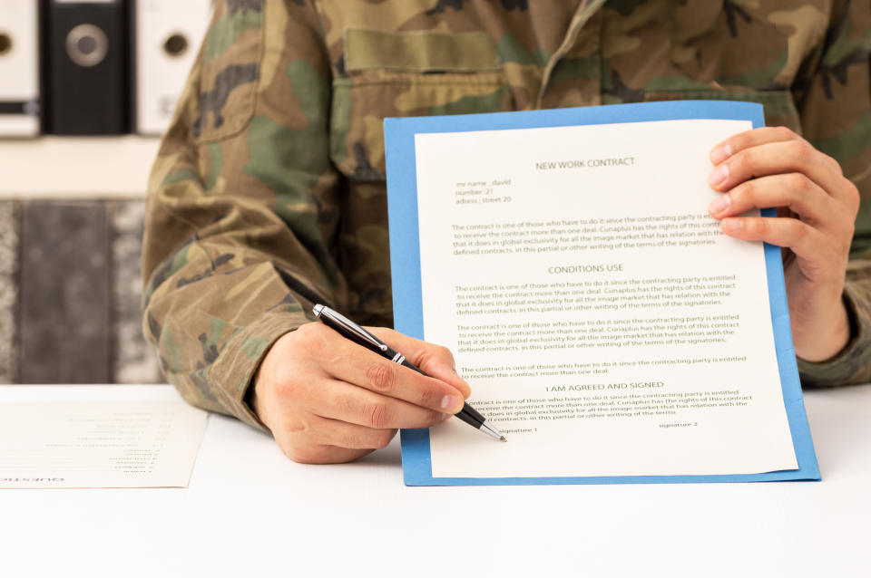 Person in military camouflage uniform holding and pointing to a new work contract document on a desk, with a pen in hand and another paper nearby