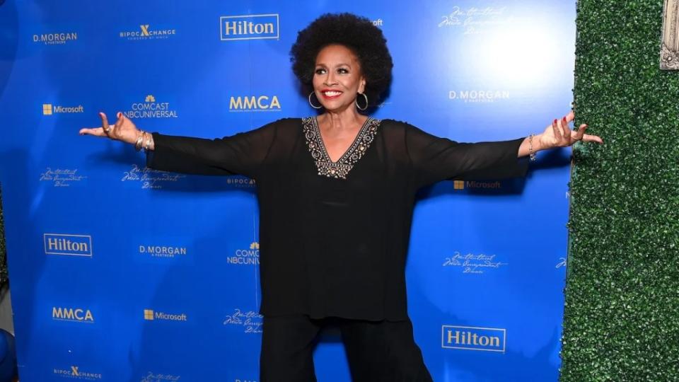 A Black woman stands with her arms outstretched in front of a blue background. It is actress Jenifer Lewis.