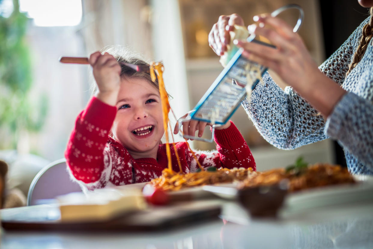 A young girl smiles widely as she holds up spaghetti bolognese on a fork while her mother grates cheese onto her plate