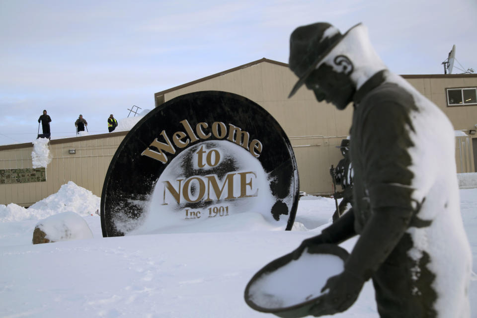 In this Feb. 14, 2019, photo, three men shovel snow from the roof of a grocery store as a statue of one of the "Three Lucky Swedes," credited with discovering gold in the late 19th century, stands in the foreground, in Nome, Alaska. The city later added statues of two native boys who led him to find the gold. (AP Photo/Wong Maye-E)