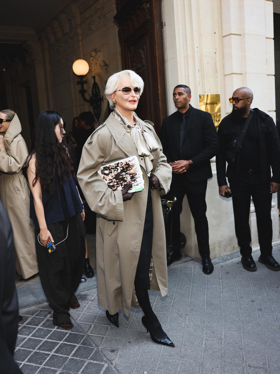 Alexis Stone walks confidently in a stylish trench coat, holding a fashion magazine. She is surrounded by security and staff
