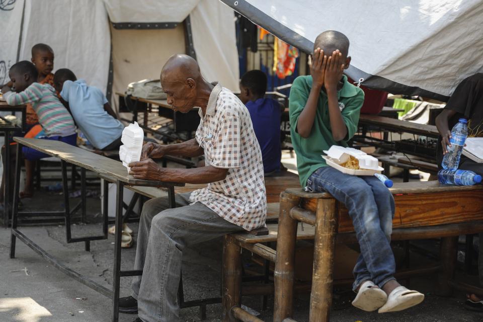 Un hombre toma su comida y un niño se cubre el rostro tras recibir envases con alimentos en un albergue para familias desplazadas por la violencia de las pandillas, en Puerto Príncipe, Haití, el 14 de marzo de 2024. (AP Foto/Odelyn Joseph)