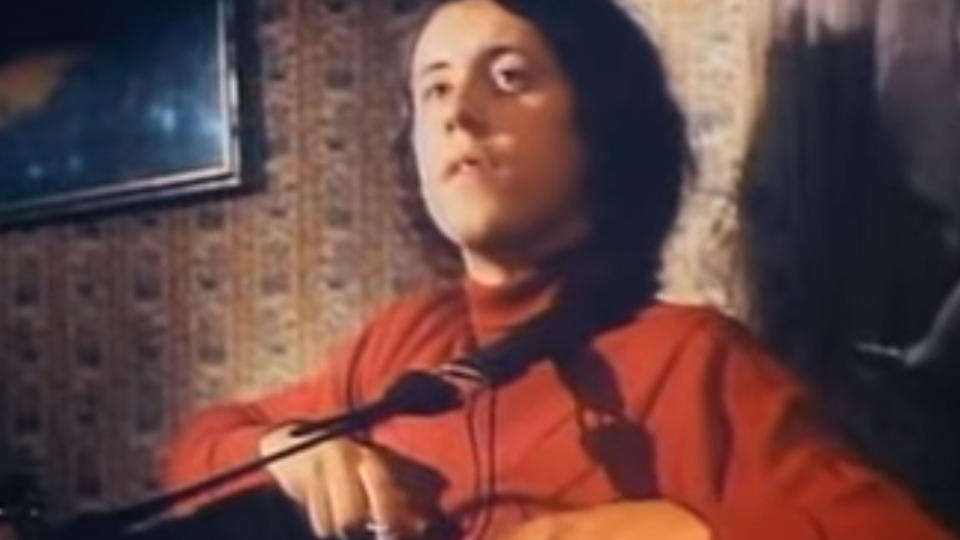 Arlo Guthrie in a red shirt, holding a guitar