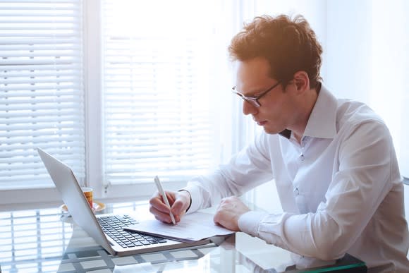 A man writes in a notebook in front of his laptop.