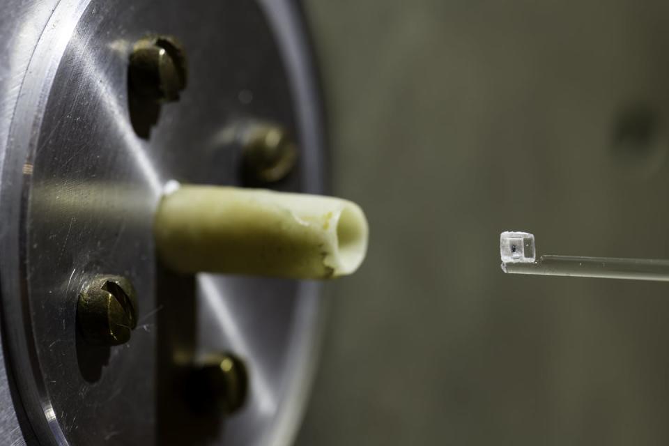 A small green fleck inside of a clear cube sitting on a stand in front of the nozzle of a scientific instrument.