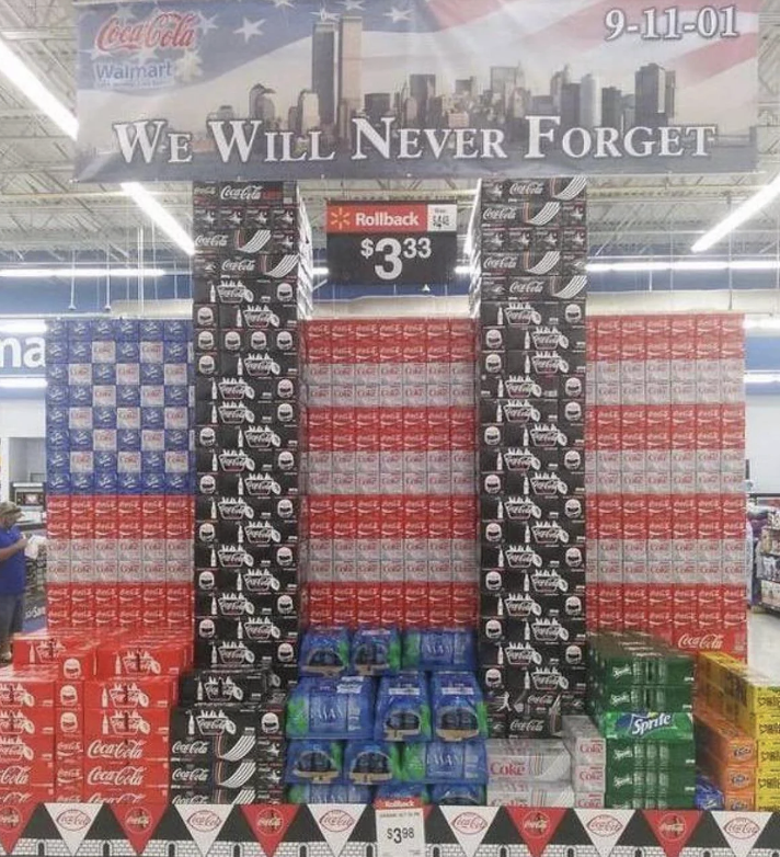 A Coca-Cola display in Walmart shows stacked soda boxes arranged in the shape of the U.S. flag with the text "We Will Never Forget" and "9-11-01" above it