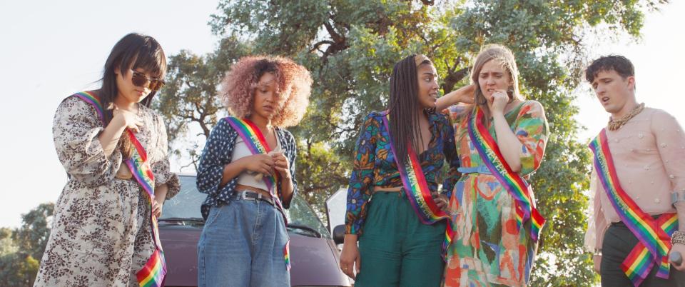 People wearing sashes with rainbow stripes stand outdoors next to a vehicle, seemingly engaged in conversation