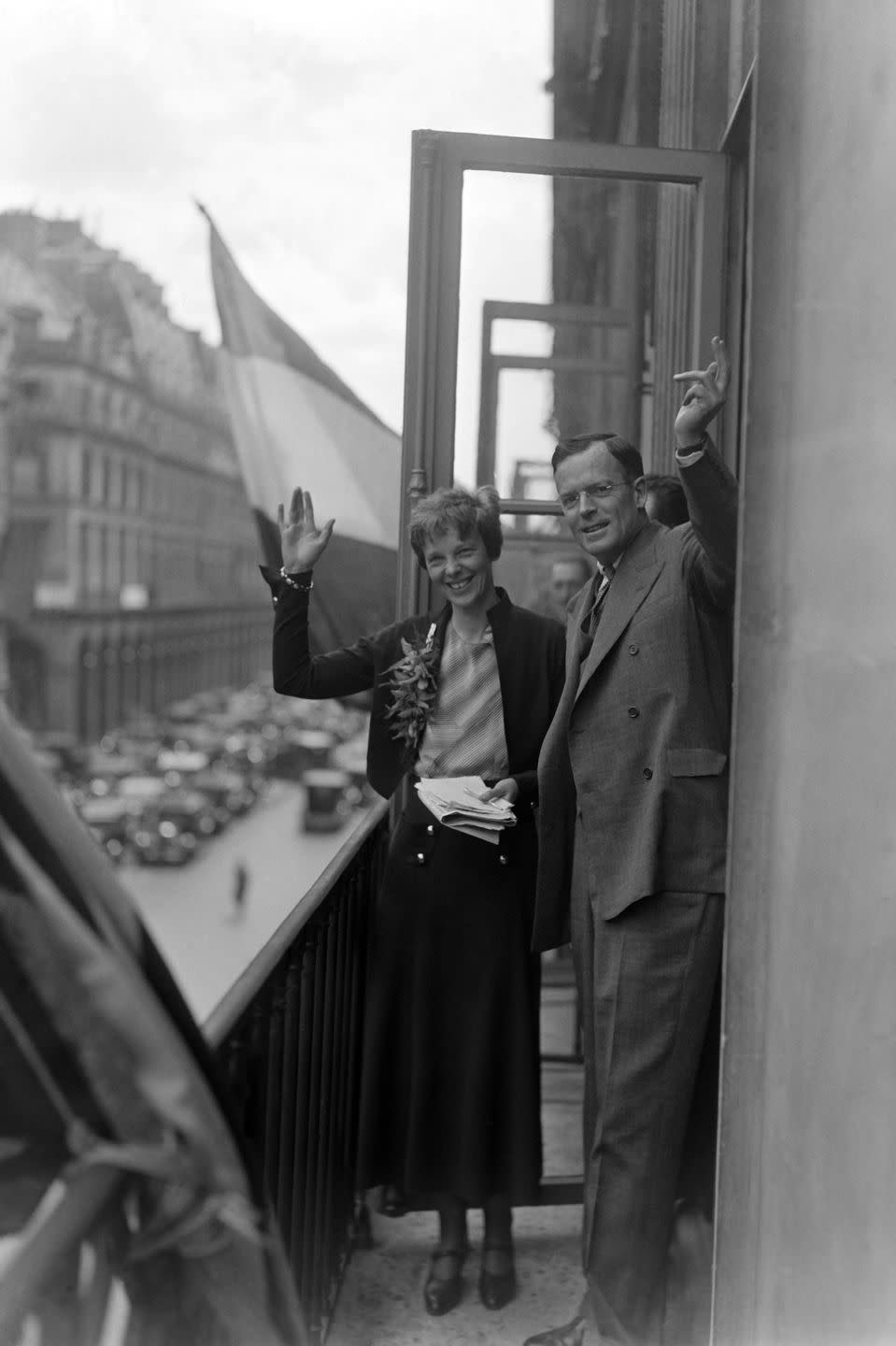 amelia earhart and george putnam wave and smile on a balcony overlooking a street, she holds a stack of papers and wears a sweater, blouse and skirt, he wears a suit