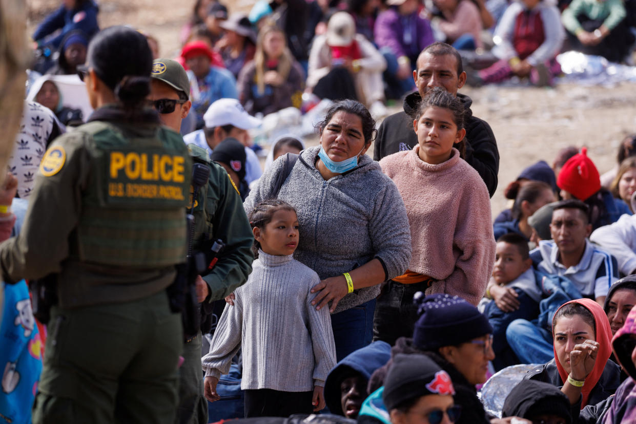 A Central American family lines up to talk to two U.S. Border Patrol agents, with many other migrants seated on the bare ground behind them.