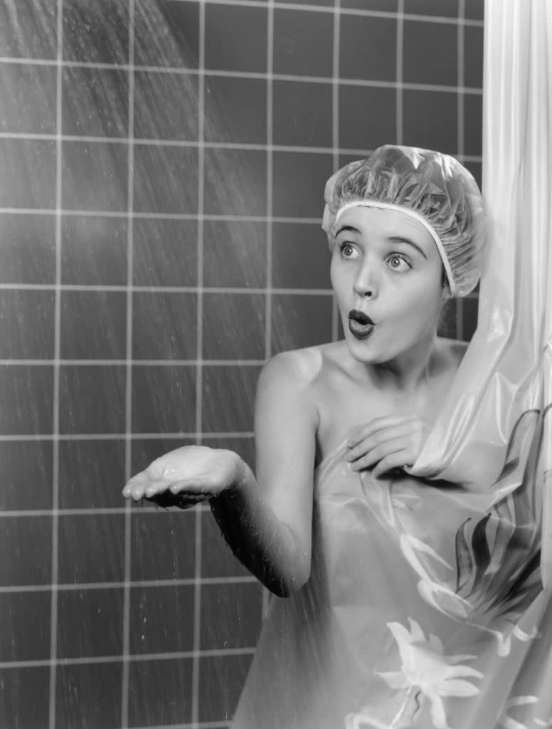 A surprised woman in a shower, wearing a shower cap and a floral shower curtain, is reaching out to feel the water. This image is related to Work & Money