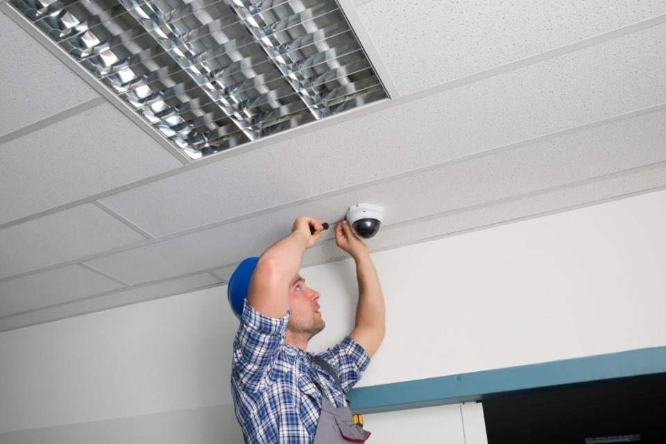 A man is seen fixing a security camera on the ceiling with a tool.