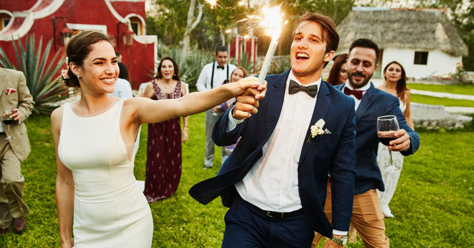 A bride and groom stand in front of their wedding party, on the grass, the groom is holding a firework