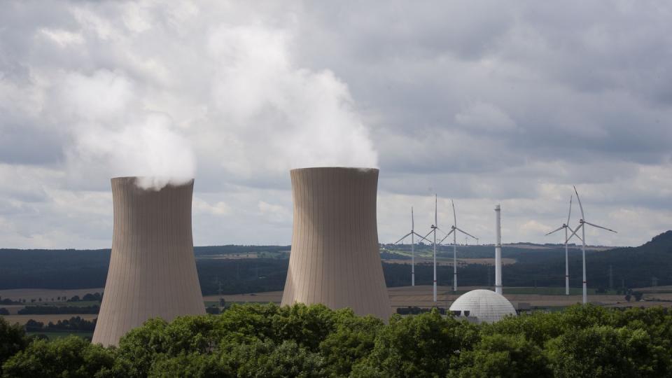 Dampf steigt aus den Kühltürmen eines  Atomkraftwerks. Im Hintergrund sind Windräder zu sehen.