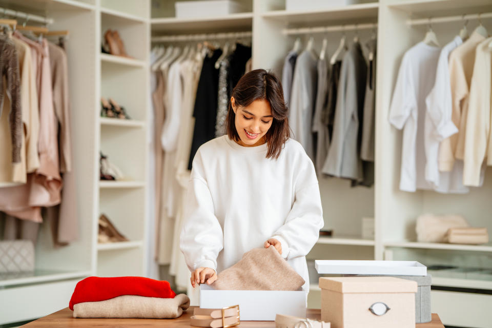 Brunette woman packing and stacking clothes items into the boxes in a big showroom or wardrobe room with a lot of clothing and shoes racks