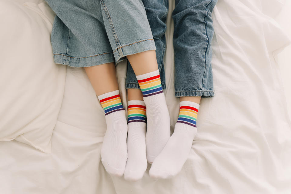Two women in jeans and socks with a rainbow print with an LGBTQ symbol lying on a bed.