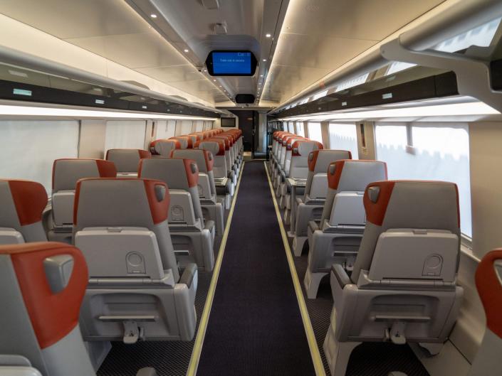 The new Amtrak Acela first class interior with rows of seats with orange headrests and a digital screen.