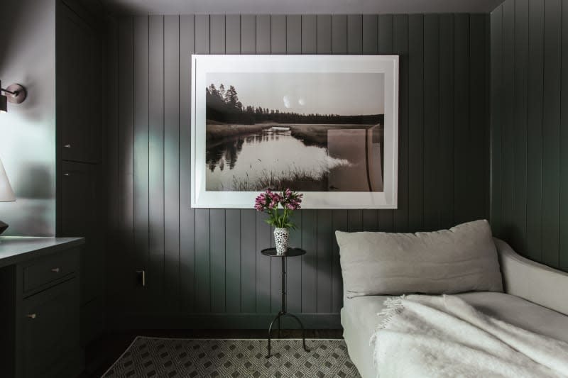 dark green tongue and groove wall paneling, pond landscape with train in background, small metal side table, small vase with flowers, low beige day bed, black and white checkered tile floor