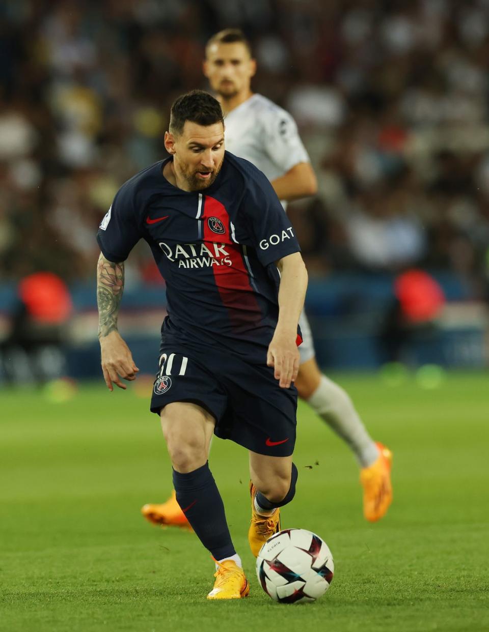 lionel messi dribbles and looks down at a soccer ball on a grass field, he wears a dark navy soccer uniform, and an opponent player watches from behind