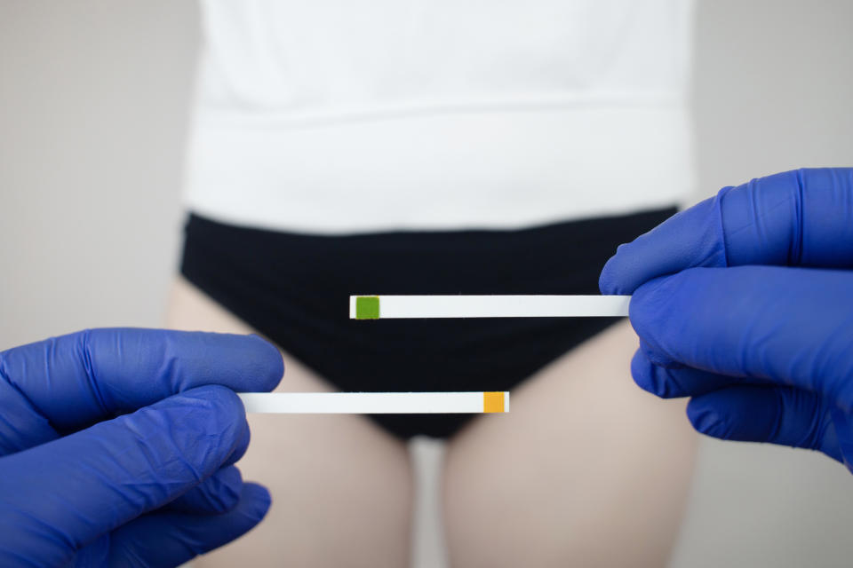 Person holding two pH testing strips, one green and one yellow, near their waist, wearing black underwear and a white top