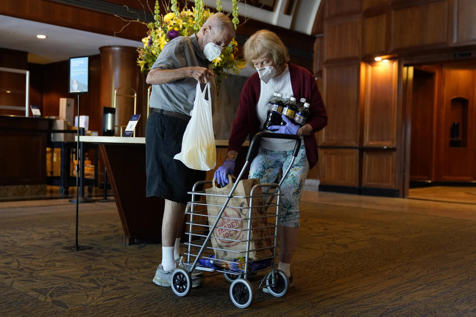 David Benkin, 85, left, and Rebecca Adler, 81, gather their grocery delivery from the lobby of their building, Wednesday Aug. 19, 2020, in North Bethesda, Md., after they were delivered by Dhruv Pai, 16, and Matthew Casertano, 15, as part of the nonprofit organization, "Teens Helping Seniors." The teens call when they are outside and leave the bags of groceries on a table in the lobby of the building for the seniors to pick up just after they leave, in order to minimize the risk of coronavirus exposure for the seniors. (AP Photo/Jacquelyn Martin)