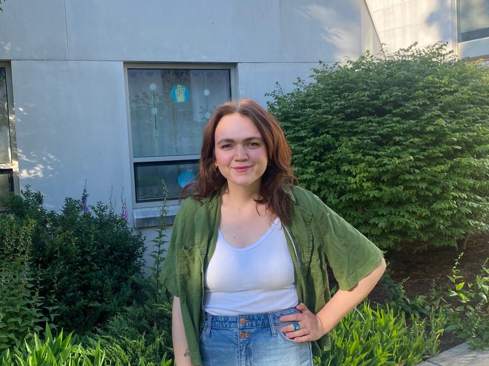 Rylie Cooper, a white nonbinary person with auburn hair, poses in front of green bushes.