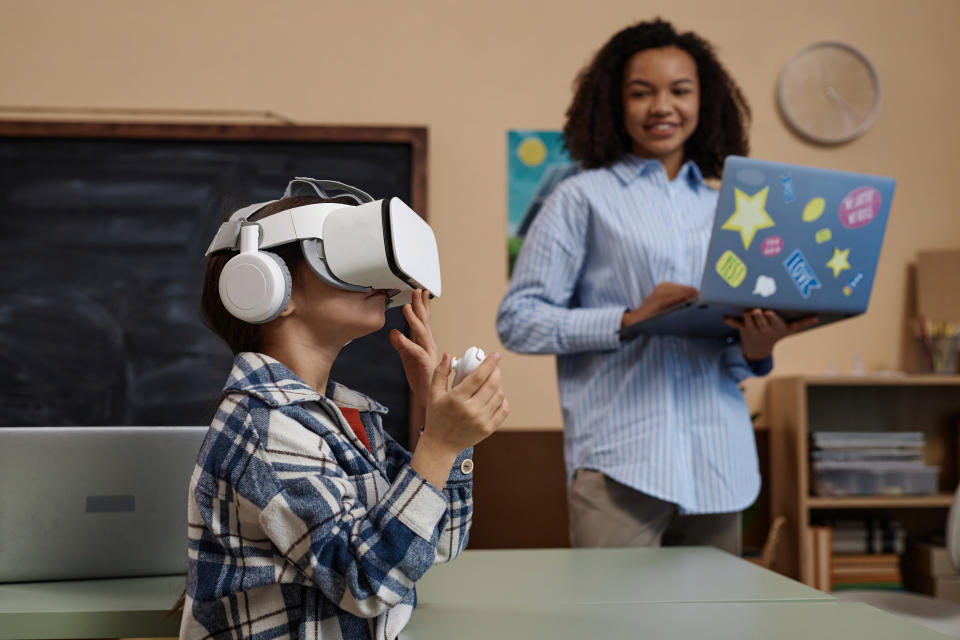 A child wearing a VR headset and a plaid shirt interacts with a virtual environment, while an adult holding a laptop watches and smiles in a classroom setting