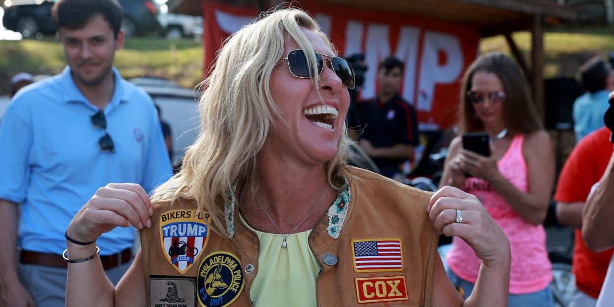 Marjorie Taylor Greene wearing a biker vest in front of a big Trump sign.