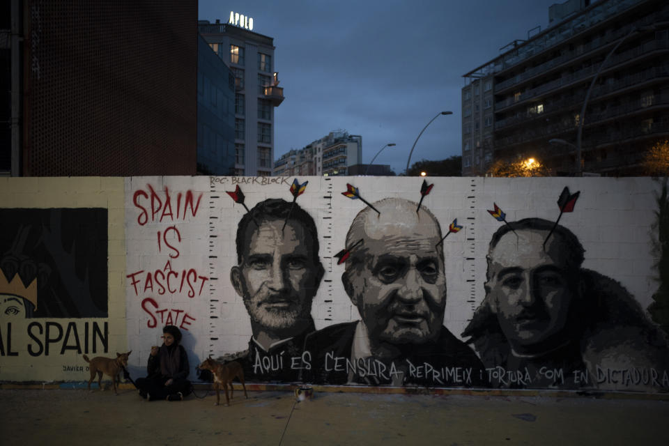 Una mujer se sienta junto a sus perros el domingo 21 de febrero de 2021 al lado de un mural que muestra, de izquierda a derecha, al rey de España Felipe VI, al exrey Juan Carlos y al fallecido dictador Francisco Franco, realizado por el artista BlackBlock, en apoyo al rapero Pablo Hasél, en Barcelona. (AP Foto/Felipe Dana)