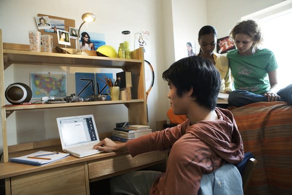 Young male at a laptop with two young females behind him who appear to be studying