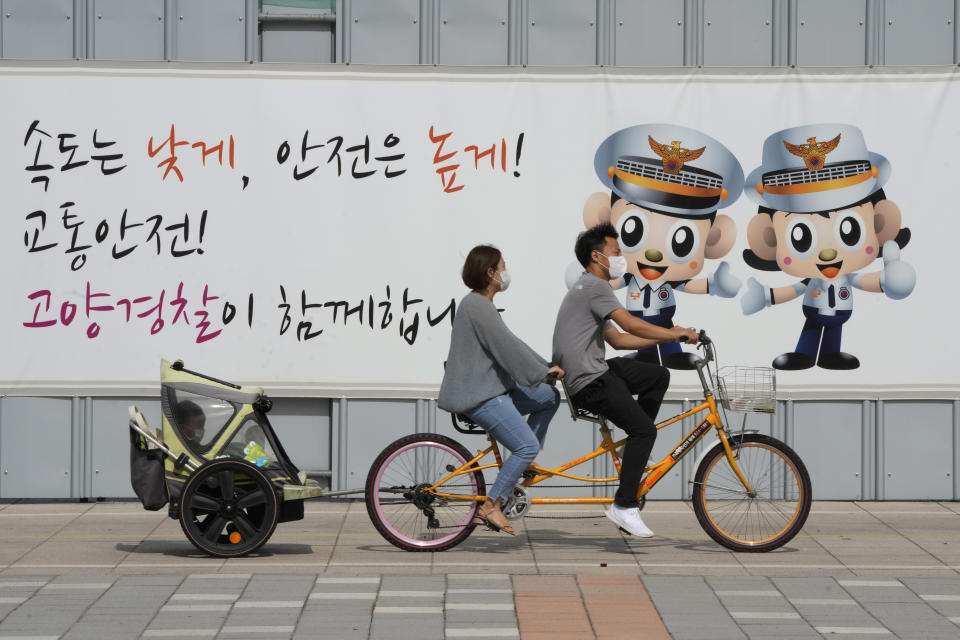 A couple wearing wearing face masks as a precaution against the coronavirus ride a bicycle with their child at a park in Goyang, South Korea, Sunday, Sept. 5, 2021. The banner reads, "Traffic safety, Goyang police are with you." (AP Photo/Ahn Young-joon)