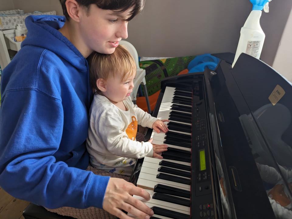 A teenager plays a piano with a baby sitting on her lap