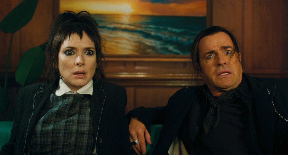 Winona Ryder and Justin Theroux look surprised as they sit on a sofa and see a painting of a sunset on the beach behind them